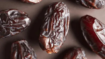 Imaginea prezintă mai multe Curmale Supreme Jumbo Medjool de dimensiuni mari, cu o textură adânc rugoasă și lucioasă, de culoare maro închis. Fiecare curmal are o formă alungită, ușor curbată, iar suprafața sa este striată, cu mici crăpături vizibile, semn al coacerii lor naturale. Aceste curmale sunt proaspete și sugerează o consistență suculentă, potrivită pentru a fi consumate ca gustare sau utilizate în diverse preparate culinare.