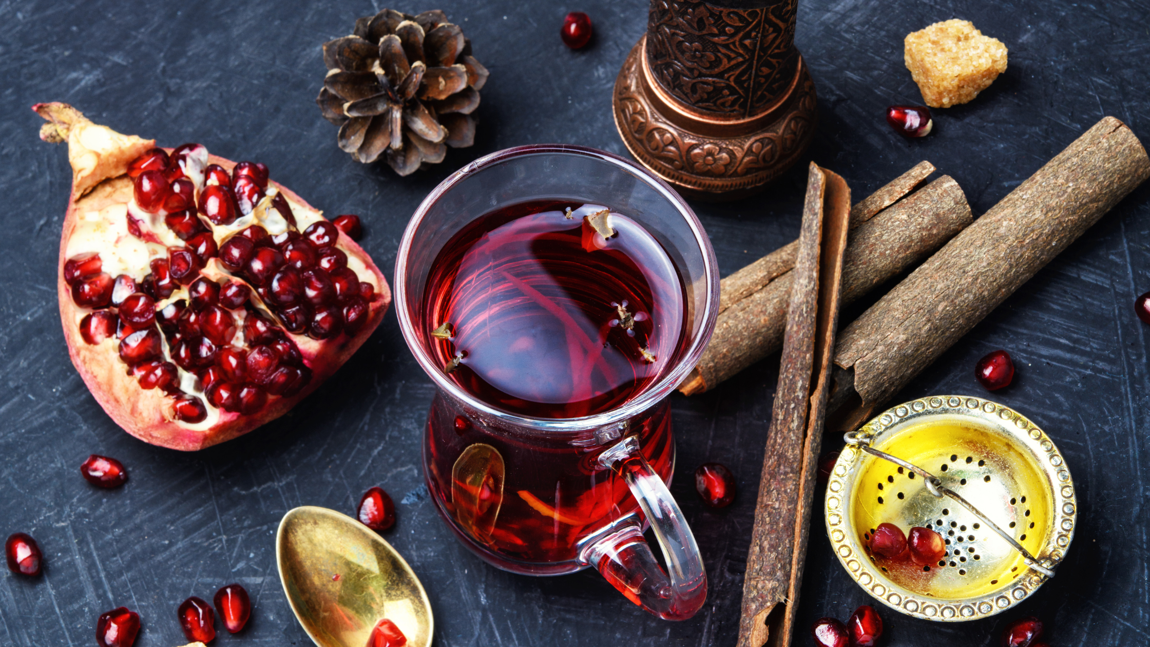 Cană de sticlă cu ceai roșu, așezată pe o suprafață închisă, alături de o jumătate de rodie proaspătă, bețe de scorțișoară, o linguriță, o sită aurie pentru infuzie și boabe de rodie împrăștiate – o compoziție naturală, caldă, de iarnă. Banner folosit pentru ilustrarea articolului ceai de rodie vs ceai de hibiscus.