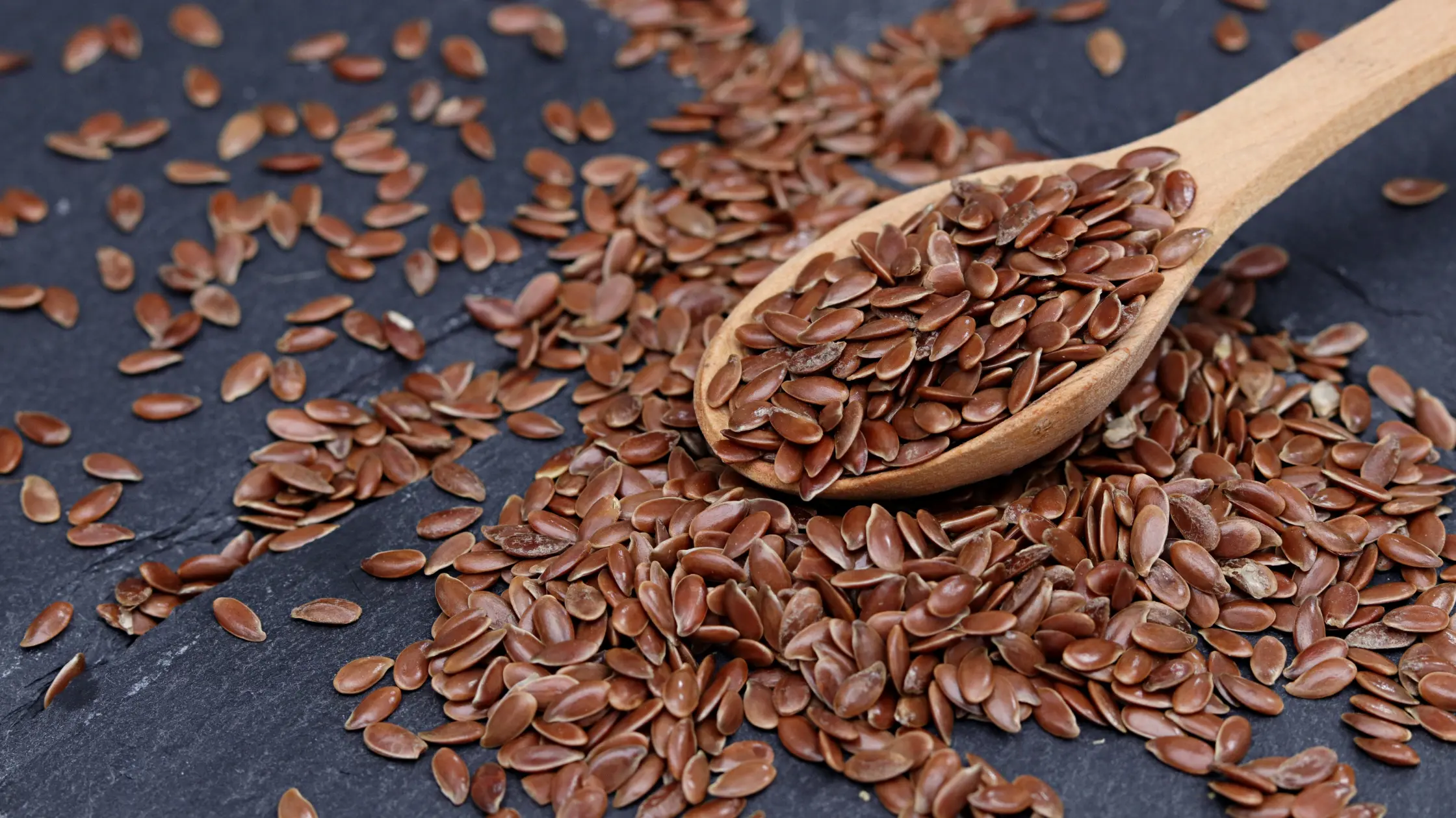 Lingură de lemn plină cu semințe de in brune, răspândite pe o suprafață de piatră închisă la culoare