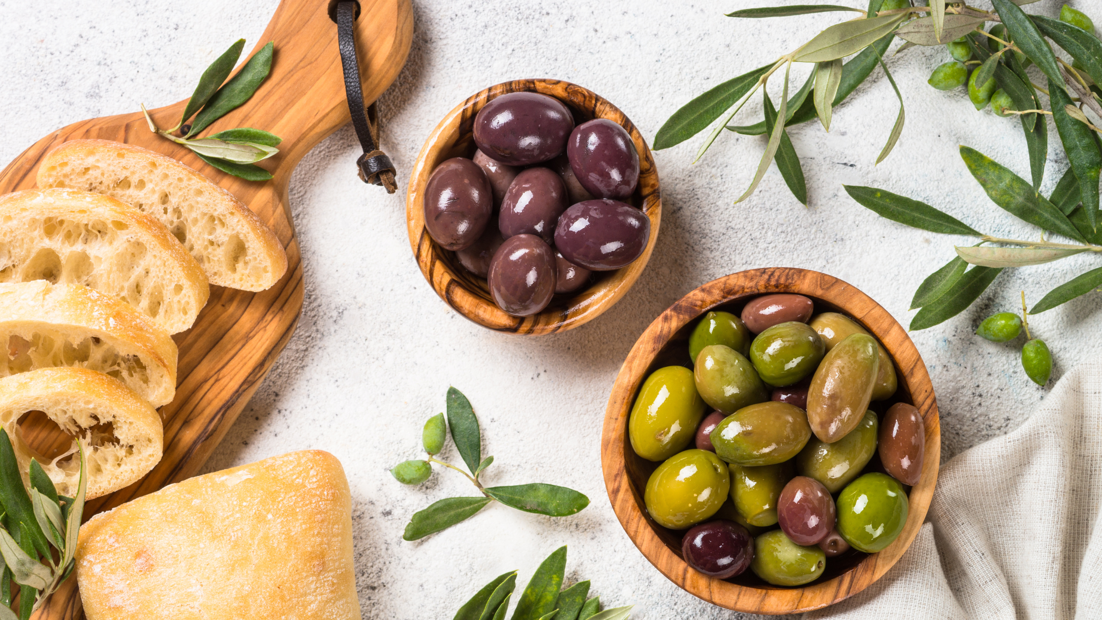 Boluri cu măsline verzi și negre, însoțite de felii de pâine rustică și frunze de măslin, așezate pe un fundal deschis – imagine care sugerează un aperitiv mediteranean savuros.