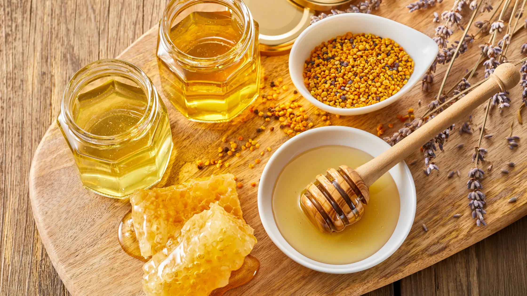 Trei borcane cu miere aurie, un bol cu polen granulat, un bol cu miere lichidă și o lingură de lemn pentru servit, așezate pe o tăblie din lemn, alături de fagure și flori de lavandă uscate – imagine care sugerează naturalețe, dulceață și idei de cadouri de Crăciun.